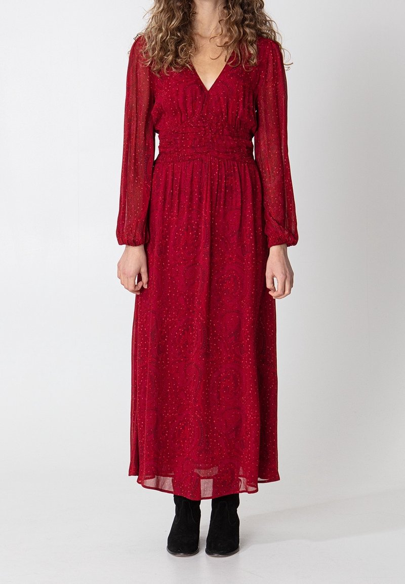 Femme portant une robe longue rouge à manches longues avec un col en V et des motifs subtils, associée à des bottines noires, debout devant un fond uni.