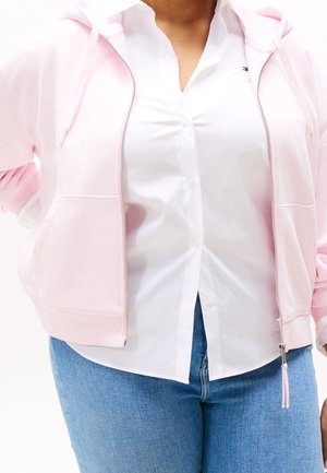 Personne portant un sweat à capuche rose clair zippé sur une chemise blanche à boutons et un jean bleu, debout les mains sur les hanches.