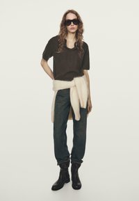 Brązowy sweter z krótkim rękawem w stylu dzianiny, kremowy pas zawiązywany w talii, ciemnoniebieskie dżinsy i brązowe kozaki za kolano. Modelka ma na sobie oversize’owe czarne okulary przeciwsłoneczne.