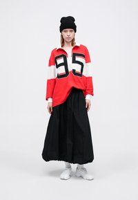 Červené a biele oversized polo s čiernym číslom "95," spojené s dlhou čiernou sukňou. Nosí sa s čiernym čiapkou a bielymi teniskami.