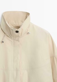 Beige jas met hoge kraag, voorzien van een ritssluiting aan de voorkant, drukknoop en verstelbare trekkoorden aan de kraag voor een betere pasvorm.