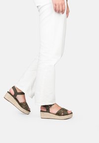 Persona con pantaloni bianchi e sandali a zeppa con stampa leopardata e cinturino alla caviglia, in piedi su uno sfondo bianco.