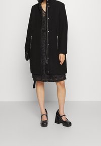 Cappotto in lana nera con chiusura a zip e cappuccio, due tasche laterali con zip. Indossato sopra un vestito in pizzo nero e abbinato a scarpe con tacco quadrato nere.