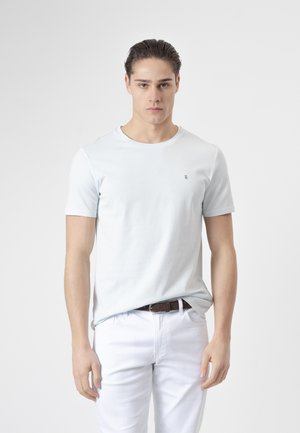 Joven con el pelo peinado hacia atrás, vestido con una camiseta lisa de color gris claro y pantalones blancos con un cinturón marrón, posando frente a un fondo blanco.