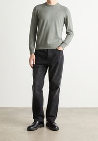 Mies, joka on pukeutunut harmaaseen pitkähihaiseen villapaitaan, mustiin farkkuihin ja mustiin nahkakenkiin, seisoo yhteen käteen taskussa harmaalla lattialla.