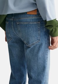 Light blue denim jeans feature a structured fit, front and back pockets, and a leather label above the back pocket. Texture appears smooth.