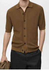 Camisa de punto marrón de manga corta con cuello, presenta un cierre de botones en la parte delantera, tela texturizada y botones oscuros. Llevada con pantalones negros.