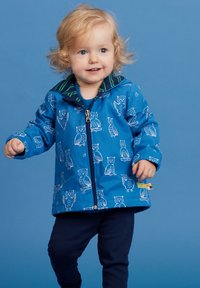 Blaue Jacke mit Eulenmustern, mit Reißverschluss und grün gestreiften Kragen. Kind trägt dunkelgraue Hosen vor einem einfarbigen hellblauen Hintergrund.