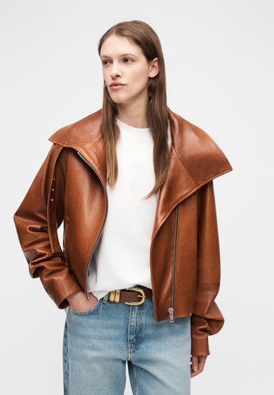Γυναίκα με μακριά καστανά μαλλιά, φοράει καφέ φαρδιά δερμάτινη τζάκετ, λευκό πουκάμισο, ανοιχτό μπλε τζιν και καφέ ζώνη, κοιτάζοντας προς τα αριστερά.