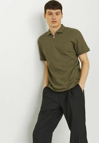 Olivgrön polo skjorta med dragkedja i kragen och korta ärmar, tillsammans med löst sittande svarta byxor. Subtilt mönster på skjortan.