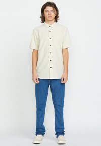 Camicia a maniche corte di un leggero beige con taschino sul petto, abbinata a jeans blu di denim e sneakers bianche, in piedi su uno sfondo semplice.