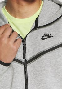 Sudadera gris con cremallera y detalles en negro, logo de Nike. Diseño con capucha, fabricada en una suave mezcla de algodón. Modelo lleva puesta una camiseta de color verde brillante debajo.