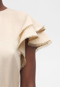 Μπεζ κοντομάνικη μπλούζα με στρωμένα φαρδιά μανίκια με λεπτή διακόσμηση κροσέ σε σκουρόχρωμο χέρι σε απλό φόντο.