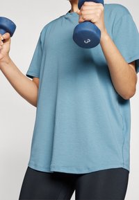 Camiseta deportiva azul hecha de tela ligera, con cuello redondo y mangas cortas, usada mientras se sostienen pesas azules.