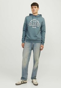 Mørk teal hoodie med en frontlomme og hvid logo-tryk. Båret med lyseblå jeans og beige sneakers med sorte striber.