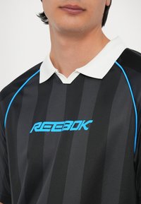Melnā un pelēkā svītrainā polo krekls ar baltu apkakli. Iespējas ir zilo akcentu un uzkrītošs zils Reebok logo uz krūtīm.
