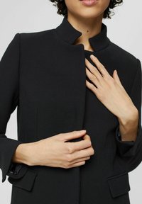 Person, der ein schwarzes maßgeschneidertes Sakko mit Stehkragen trägt, berührt mit einer Hand in der Nähe der Brust das Sakko und mit der anderen an der Taille.