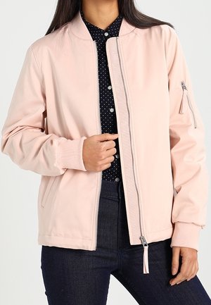 Vrouw draagt een lichtroze bomberjack over een donkerblauw blouse met witte stippen en donkere spijkerbroek, houdt met één hand de rits van het jack vast.