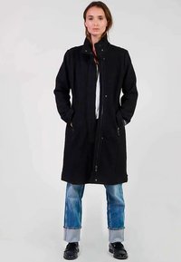 Lange zwarte jas met een hoge kraag, ritssluiting aan de voorkant en zijzakken, gecombineerd met een blauwe spijkerbroek en zwarte schoenen. Gladde textuur en minimalistisch ontwerp.