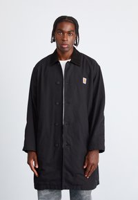 Sort frakke med blød krave, forreste knapper og Carhartt-logo patch. Har en afslappet pasform og lange ærmer. Tekstureret stof.
