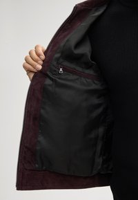Giacca in suede bordeaux con fodera interna in raso nero; presenta tasca interna con zip e chiusura con zip in metallo.