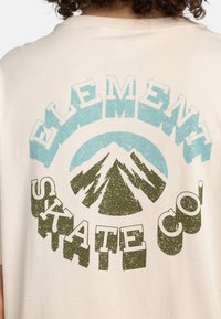 Bežové bavlněné tričko s grafickým potiskem, který zobrazuje modré, zelené a černé hory, a s výrazným nápisem: "ELEMENT SKATE CO."