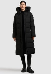 Geacă puffer neagră cu un design lung și supradimensionat, având o culoare înaltă, un model cu matlasare orizontală și fermoar frontal. Asortată cu cizme negre.