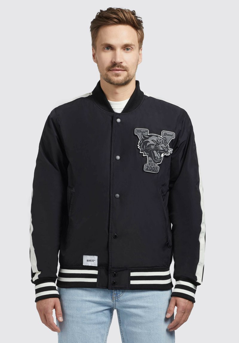 Zwarte bomberjack met contrasterende witte strepen op de mouwen, een panterlogo patch en ribboorden aan de mouwen en onderkant. Gemaakt van gladde stof.