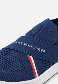 Marinblå sneakers med texturerad mesh-övre, elastiskt band med vita och röda ränder, och vit gummisula. Varumärkeslogotypen visas framträdande.