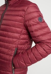 Rote Steppjacke mit horizontalem Steppmuster, Reißverschlusstasche und rundem Logodetail am linken Ärmel. Glatte, glänzende Textur.