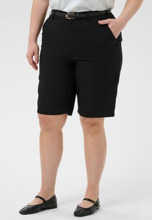 Kvinde iført sorte knælange skræddersyede shorts med sort bælte, hvid top og sorte flade sko, stående med den ene hånd i lommen.
