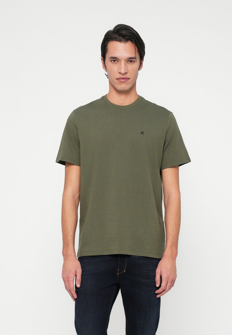 Mann står iført en olivengrønn t-skjorte med korte ermer og liten svart logo på brystet, samt mørkeblå jeans, mot en ensfarget hvit bakgrunn.