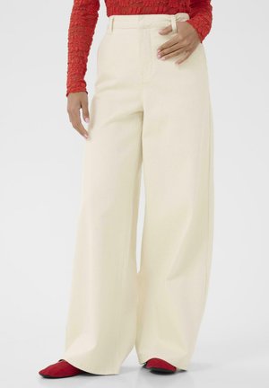 Žena oblečená v krémových širokých nohaviciach s vysokým pásom, červenom textúrovanom dlhorukávovom topu a červených špicatých balerínach, ruka opretá o pás.