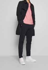 Czarny płaszcz trencz, różowy t-shirt i ciemne spodnie noszone z białymi trampkami. Charakteryzuje się smukłym krojem i standardowym dopasowaniem, z ozdobnymi guzikami.