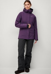 Lila wasserdichte Jacke mit Kapuze, Reißverschluss und Seitentaschen, kombiniert mit schwarzen Hosen und dunklen Schuhen. Glatte Textur, Outdoor-Design.