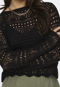 Pull noir en tricot avec un motif ajouré, présentant un ourlet festonné et des manches longues. Accentué par un collier chaîne dorée.