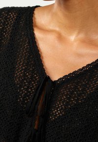 Czarny szydełkowany top z dekoltem w kształcie V, teksturowanym wzorem i ozdobnymi sznurkami z przodu. Miękki, lekki materiał.