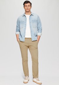  Lichtblauw denim shirt met twee borstzakken, wit T-shirt, beige broeken, beige sneakers met crème accenten, staande pose tegen een effen achtergrond.