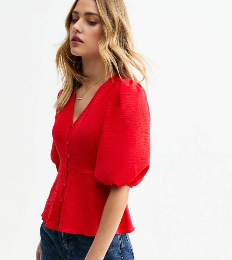 Blouse rouge à manches bouffantes avec un décolleté en V profond, tissu texturé, fermeture à boutons sur le devant et taille ajustée, associée à un jean en denim bleu.