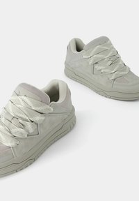 Coppia di scarpe sportive grigio chiaro con suole spesse e strutturate e lacci larghi e decorati su sfondo bianco semplice.