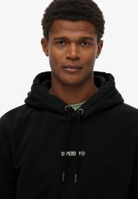 Sudadera de forro polar negra con cordón ajustable, que presenta un logo en texto blanco. El diseño incluye una textura suave y un corte relajado.