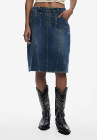 Gonna a tubino di jeans con una forma aderente, lavaggio blu scuro, tasche frontali e cuciture decorative. Abbinata a stivali da cowboy neri con dettagli bianchi.