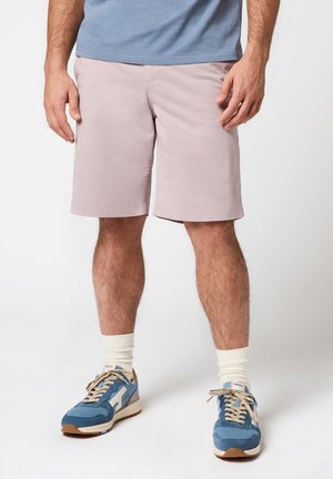Homme portant un short rose clair, des baskets bleues avec des lacets beiges, des chaussettes blanches et un t-shirt bleu, debout devant un fond blanc.