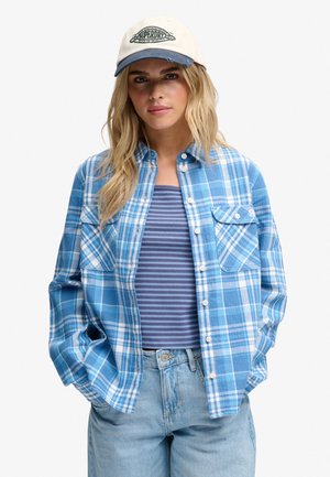 Junge Frau mit langen blonden Haaren, die eine weiße Mütze, ein blau kariertes Hemd mit Knöpfen, ein blau gestreiftes Oberteil und hellblaue Jeans trägt und die Hände in den Taschen hat.