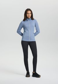 Hellblaue Zip-Jacke mit strukturiertem Muster, hohem Kragen und zwei Seitentaschen, kombiniert mit schwarzen enganliegenden Leggings und schwarzen Sneakers.