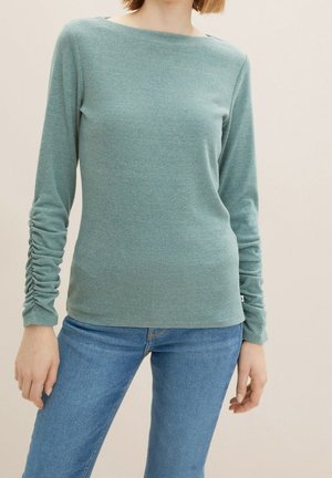 Person, die ein blau-grünes Langarmoberteil mit gerafften Ärmeln und blaue Jeans trägt, steht vor einem einfarbigen hellen Hintergrund.