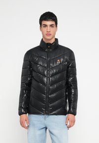 Juoda pūsta striukė su aukštu apykakliu, chevrono raštu, užtrauktuko uždarymu ir nedideliu logotipu ant krūtinės. Lygus, blizgus paviršius.