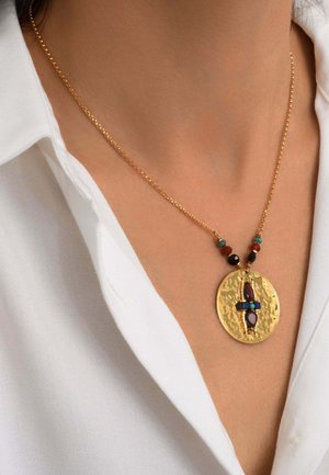 Collier chaîne en or avec un grand pendentif rond martelé orné de pierres multicolores, porté sur un col de chemise blanche ouverte.