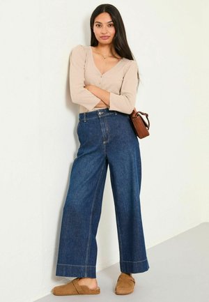 Wide-leg dark blue denim jeans with a high waist, paired with a light beige long-sleeve top and tan slip-on shoes. Small brown handbag.
