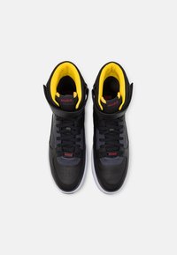 Höga sneakers i svart syntetmaterial med gul foder. Funktioner inkluderar snörning, perforerat tåområde och röda varumärkesaccenter.